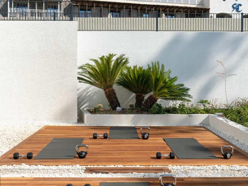 Terrasse en bois avec tapis de yoga et haltères, mur blanc et palmiers en arrière-plan.