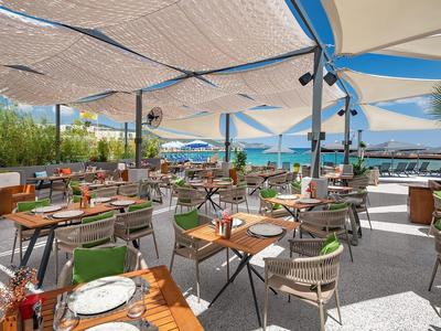 Open-air restaurant with modern wooden tables and chairs, sunshade, and sea view.