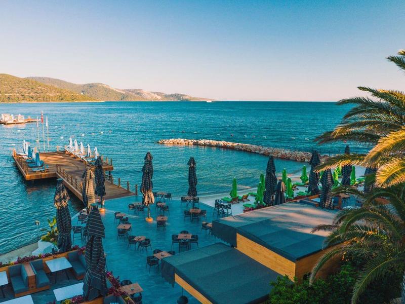 Resort de playa con piscina, palmeras y vista al mar azul y montañas al fondo.