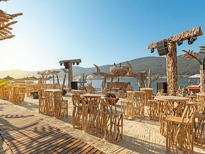 Sunny beach bar with wooden chairs and tables in front of hills under blue sky.