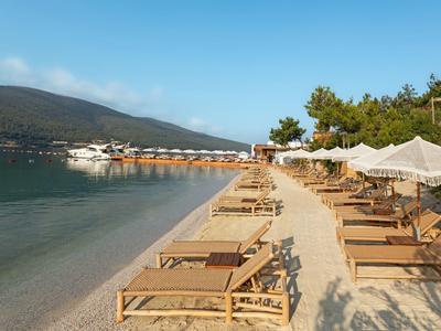 Empty beach with loungers and umbrellas next to calm water and wooded hills.