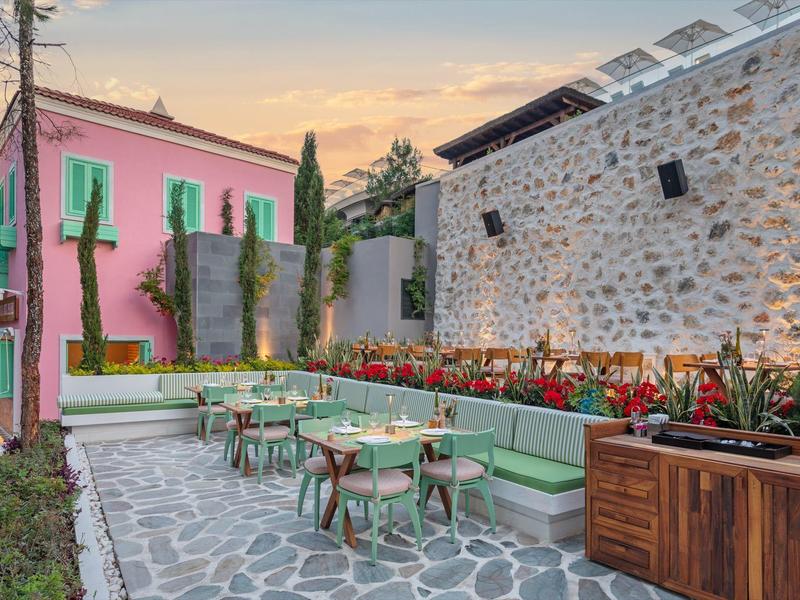 Colorful terrace with tables, chairs, and a long bench next to a stone facade at sunset.