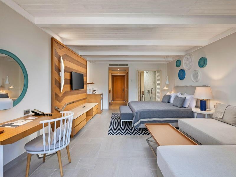 Modern hotel suite with double bed, sofa, desk, and flat-screen TV in a bright setting.