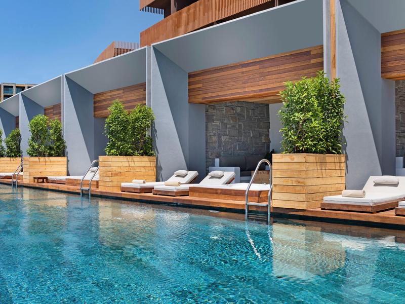 Modern hotel pool with sun loungers and private niches, surrounded by wood and plants.