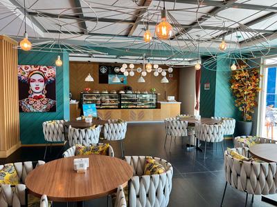 Modernes Café mit Holztischen, grauen gepolsterten Stühlen und Kunst an den Wänden bei Tageslicht.