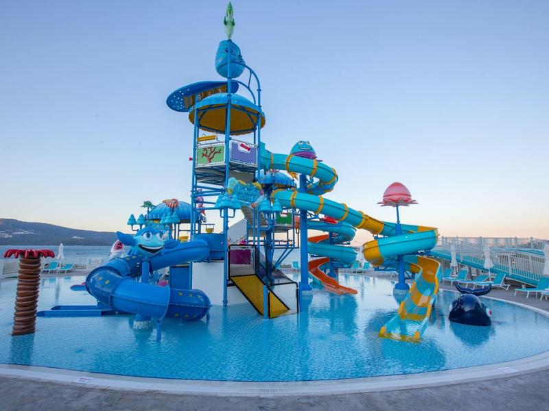 Vodní park s modrými a žlutými skluzavkami obklopený bazénem pod jasnou oblohou.