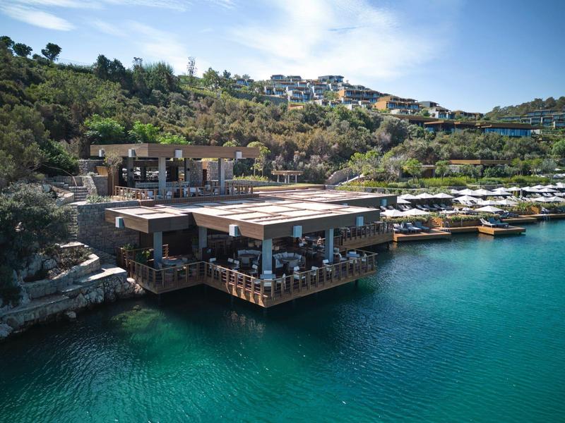 Restaurante moderno frente al agua con vista a una colina verde y cielo soleado.