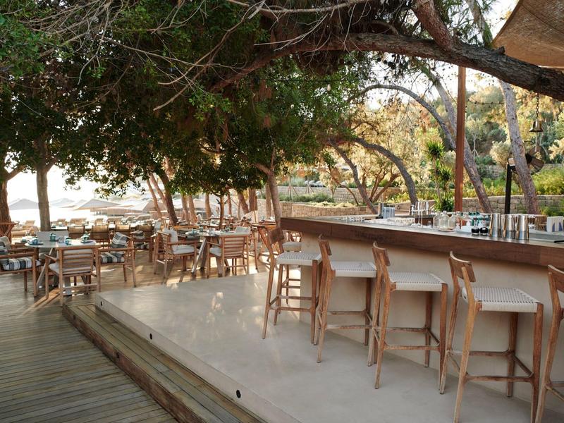 Área de bar al aire libre con taburetes de madera y zona de comedor sombreada bajo árboles junto al agua.