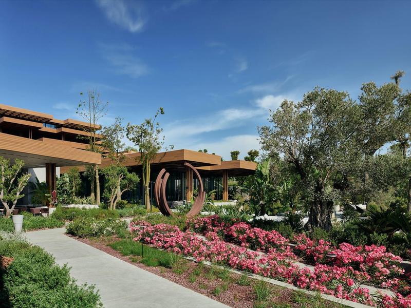Un jardín bien cuidado con flores, árboles y edificios modernos bajo un cielo azul.