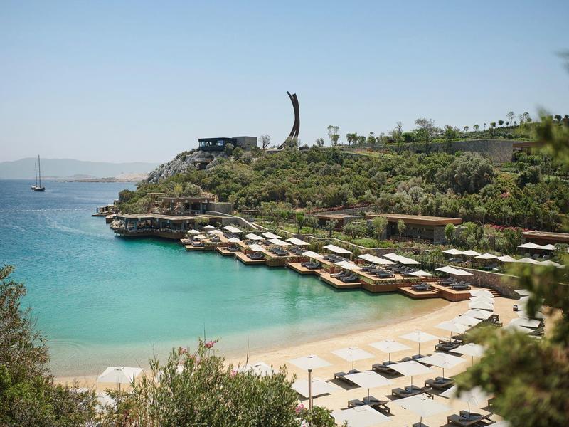 Playa con tumbonas y sombrillas junto a aguas claras y colinas verdes bajo un cielo despejado