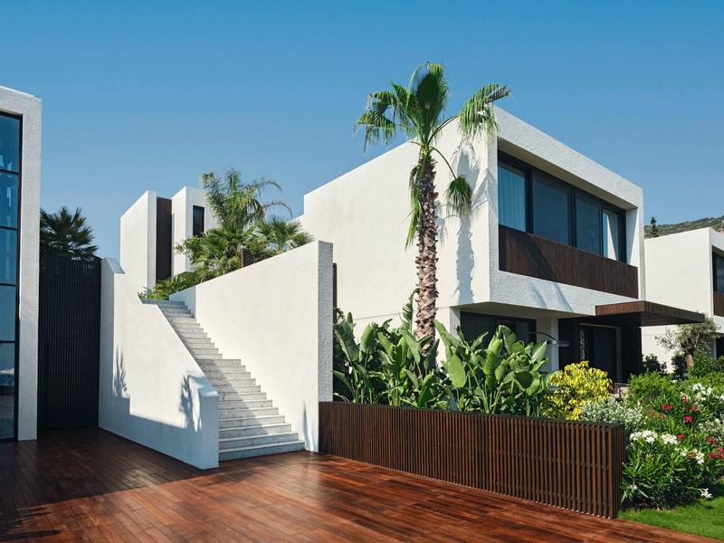 Villa blanca moderna con jardín, terraza y palmeras bajo cielo azul despejado.