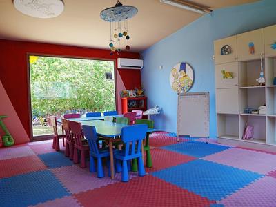 Kolorowy pokój zabaw dla dzieci z stołami, krzesłami i dużymi oknami z widokiem na zieleń.