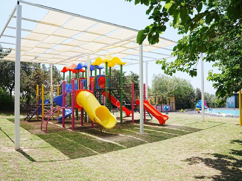 Kolorowy plac zabaw z zjeżdżalniami i konstrukcjami do wspinaczki pod białym daszkiem przy basenie.