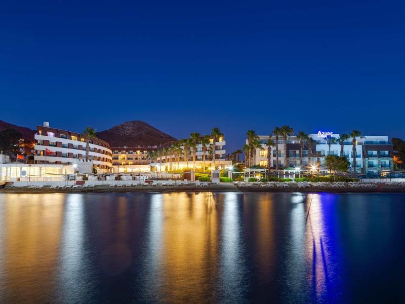 Illuminated hotel buildings by the water with mountain backdrop at night.