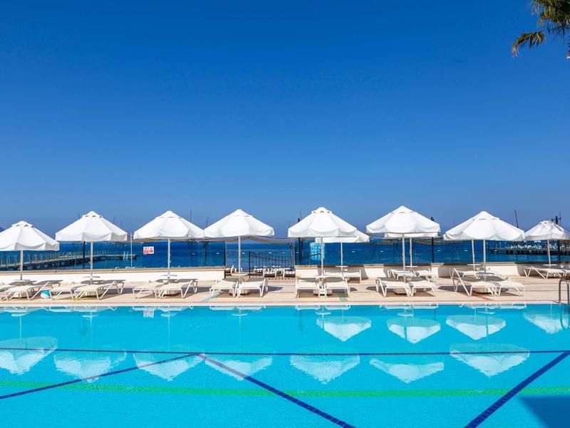 View of a hotel pool with white umbrellas and loungers by the sea under clear skies.