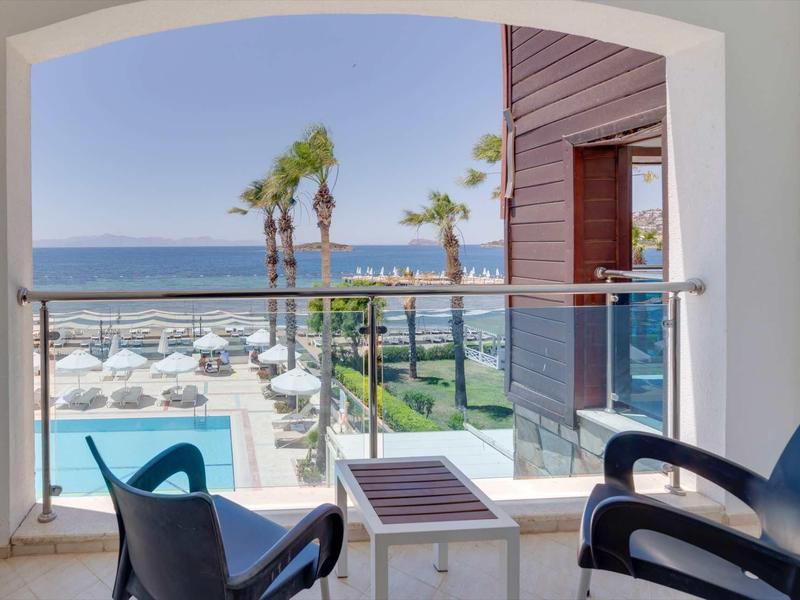 View from a balcony with two chairs overlooking a pool, palm trees, and the sea under clear sky.