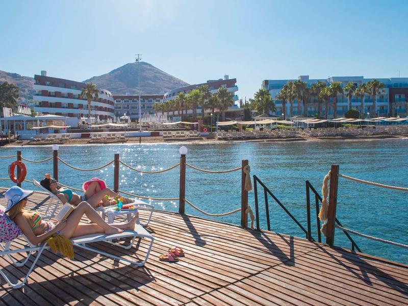 Person sunbathing on a lounge chair on a pier overlooking water and buildings.
