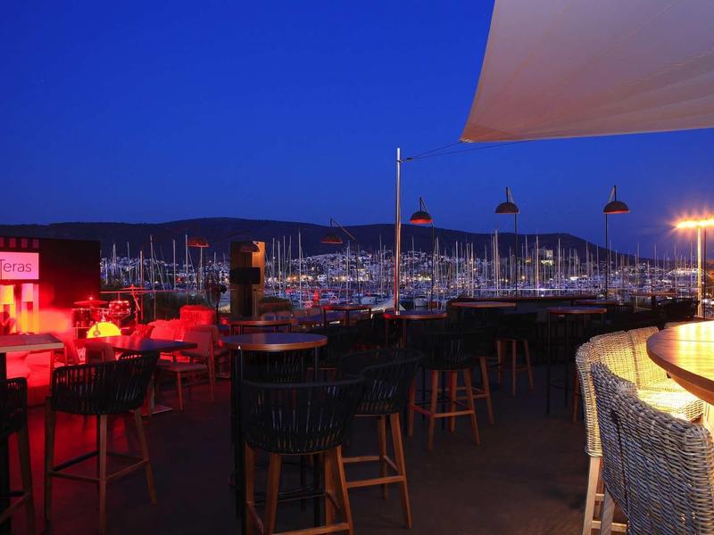 Vista notturna di un bar con vista su un porto turistico pieno di barche a vela illuminate.
