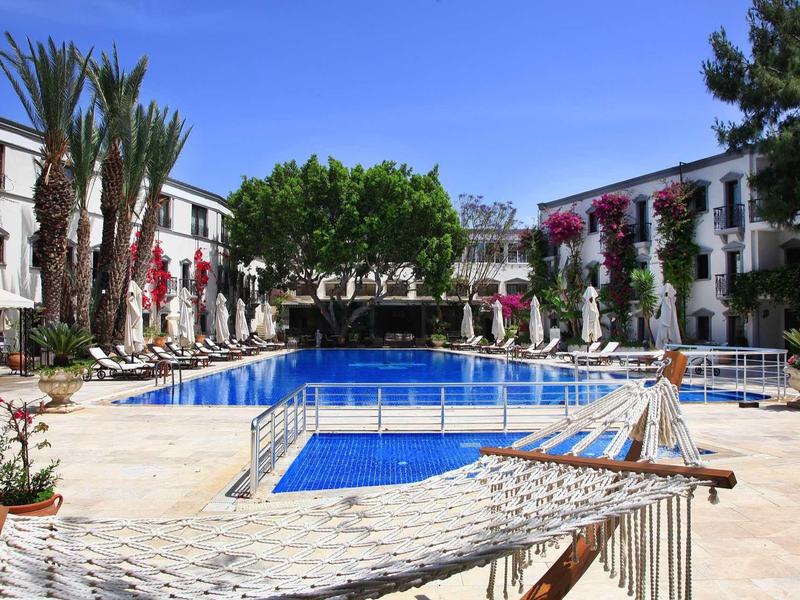 Piscina dell'hotel con acqua blu, circondata da edifici bianchi e lettini sotto un cielo blu.