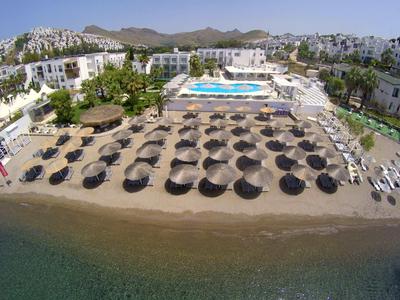 Plage de sable avec parasols devant un hôtel avec piscine et montagnes en arrière-plan.