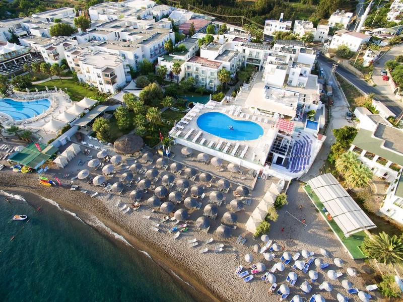 Vista aerea di un resort sulla spiaggia con piscine, ombrelloni e edifici vicini.
