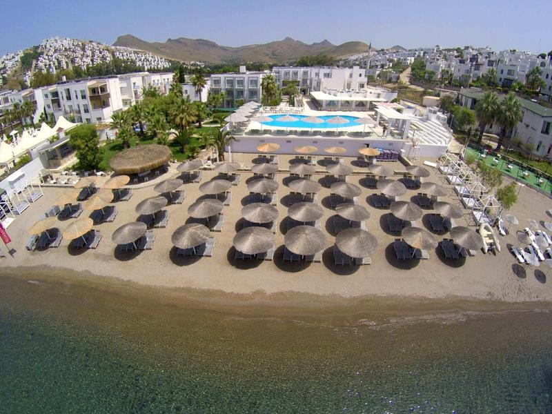Hotel sulla spiaggia con piscina, lettini e ombrelloni vicino al mare, circondato da edifici bianchi.