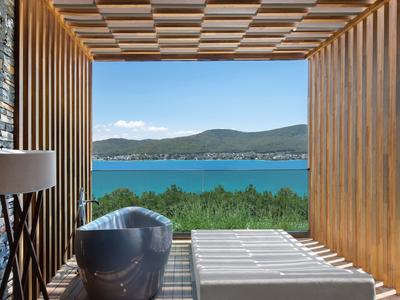 Modern wooden terrace with bathtub overlooking a blue bay and green hills.