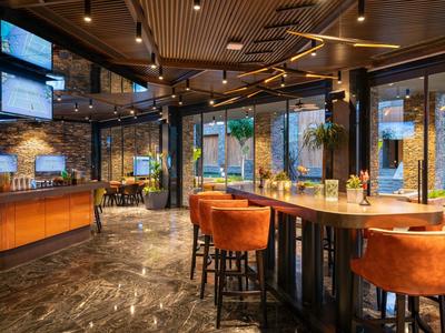 Modern hotel bar with bright chairs, wooden tables, and large windows.