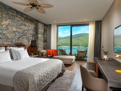 Modern hotel room with stone wall, large bed, sofa, and mountain view through floor-to-ceiling windows.