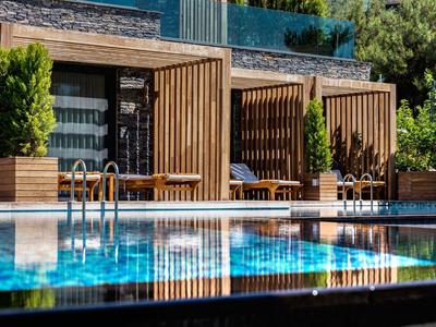 Modern wooden cabanas with sun loungers beside a clear outdoor pool in a green setting.