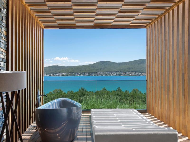 Modern wooden terrace with bathtub overlooking a blue bay and green hills.