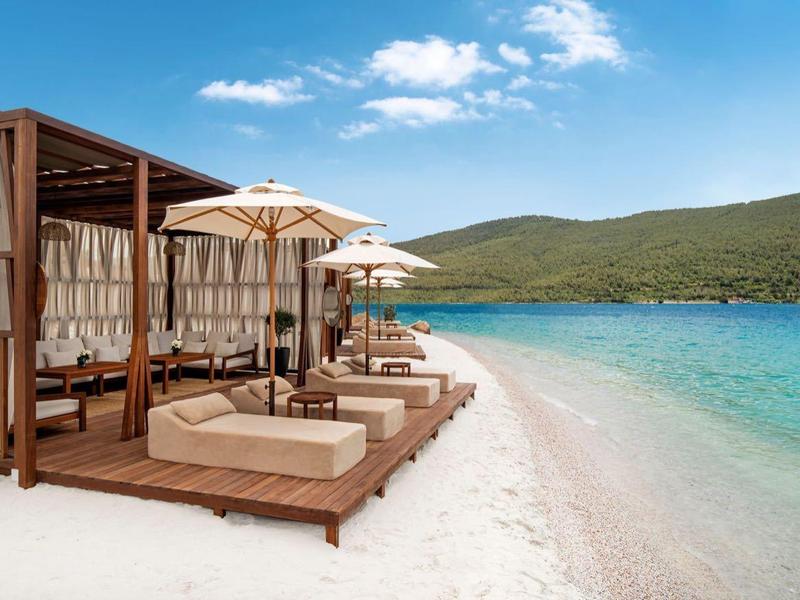 Beach with cozy daybeds and umbrellas by clear turquoise water and green hills.