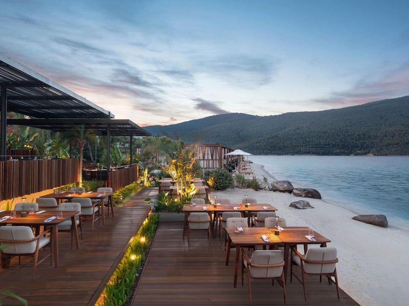 Terrace with tables and chairs by the water with hills in the background at sunset.