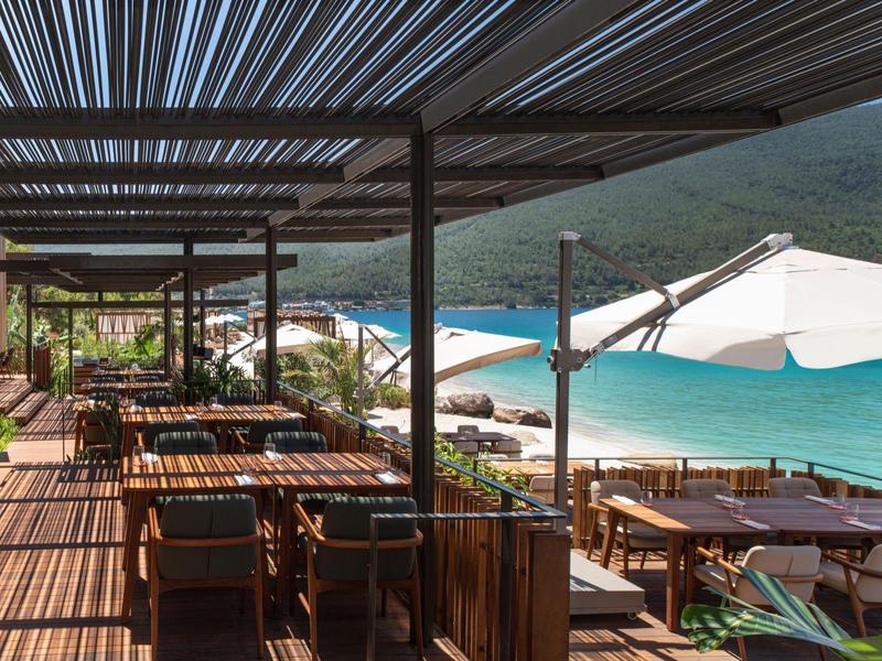 Open terrace with tables and chairs next to sandy beach and blue sea under sun umbrellas.