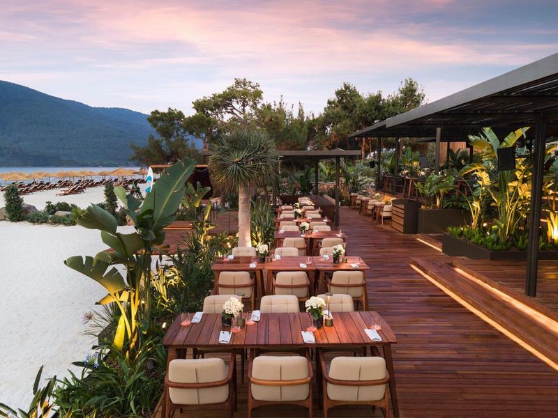 Hotel terrace with wooden tables and cushioned chairs, overlooking water and mountains.