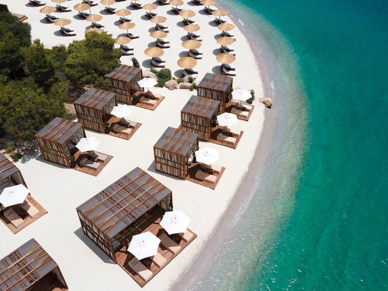 Beach with wooden cabanas, white umbrellas, and turquoise sea beside a wooded area.