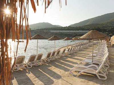 Row of lounge chairs with umbrellas by calm water and mountains in the background at sunset.
