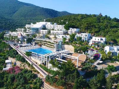 Hotel complex with white buildings and large pool in green, hilly landscape area.