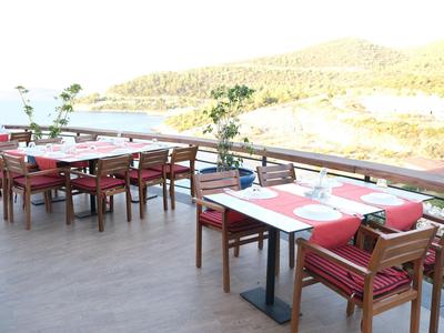 Espace de restauration extérieur avec tables en bois, chaises rouges et vue sur l'eau et les collines.