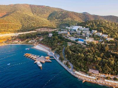 Station balnéaire côtier avec marina, plages, collines verdoyantes et bâtiments dispersés sous un ciel clair.