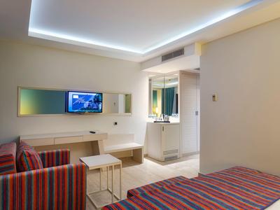 Chambre d'hôtel moderne avec lit et canapé rayés, petites tables, plafond éclairé et TV murale.