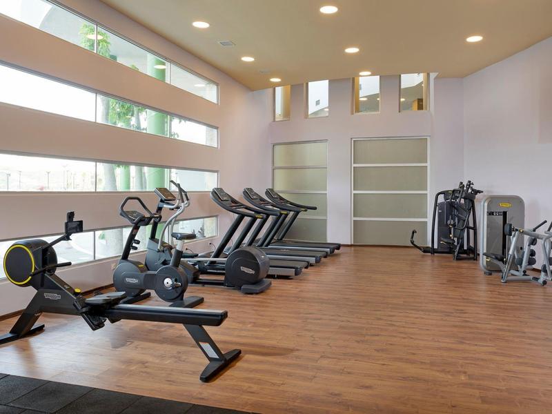 Salle de sport lumineuse avec sol en bois, tapis de course, rameurs et appareils de musculation.