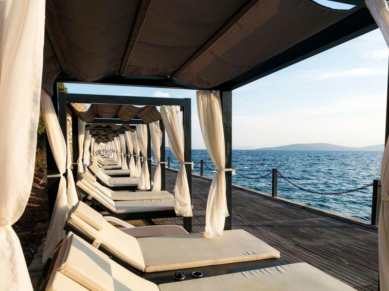 Alignement de lits de plage blancs avec rideaux sur une jetée en bois au bord de la mer au coucher du soleil.
