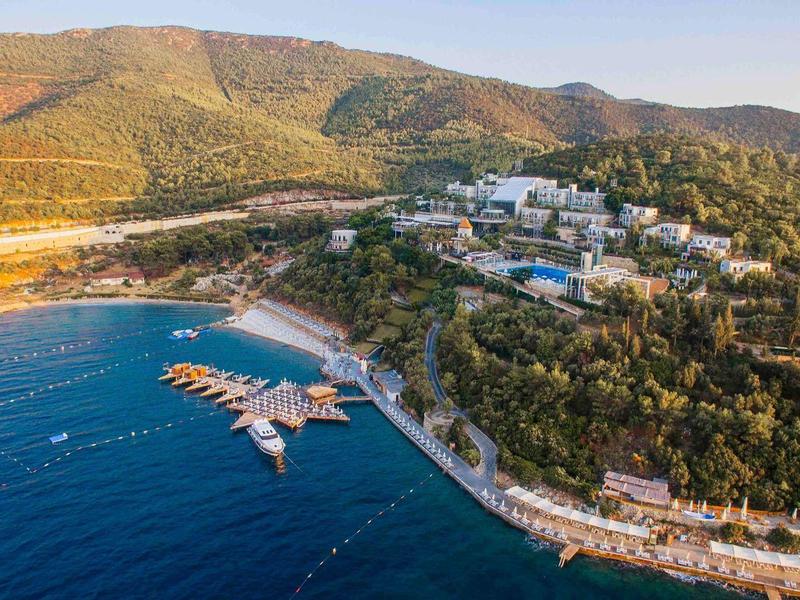 Station balnéaire côtier avec marina, plages, collines verdoyantes et bâtiments dispersés sous un ciel clair.