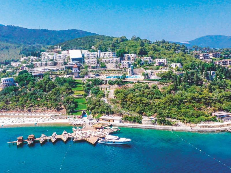 Station balnéaire côtier avec plage, jetée et bâtiments sur une colline sous un ciel clair.