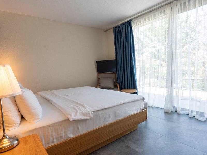 Chambre d'hôtel lumineuse avec grand lit, lampe de chevet et fenêtre du sol au plafond avec rideaux.