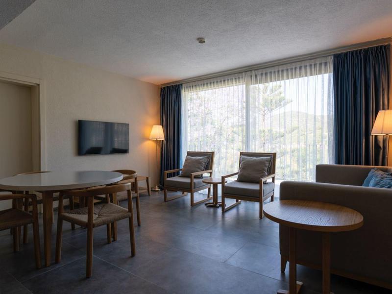 Chambre d'hôtel moderne avec table à manger, chaises, canapé et grande fenêtre avec rideaux.
