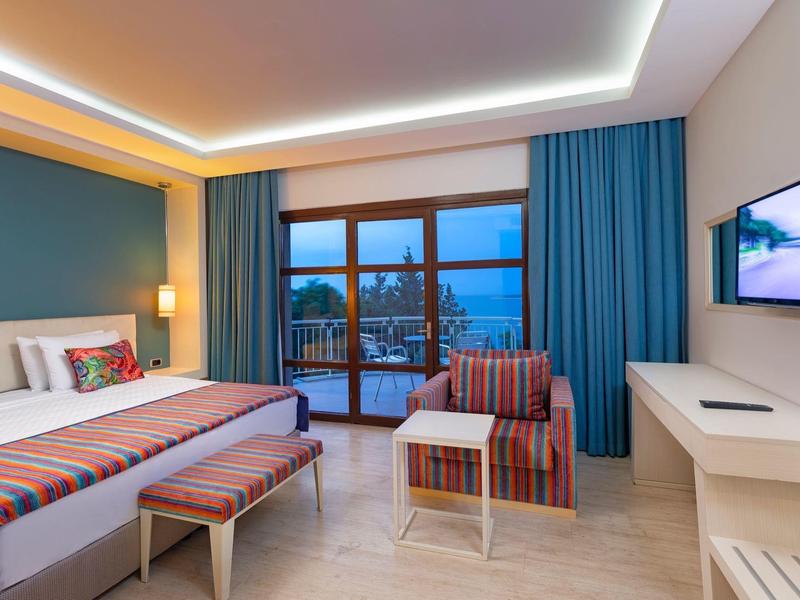 Chambre d'hôtel avec deux lits, banc rayé, fauteuil et balcon avec vue sur mer au crépuscule.