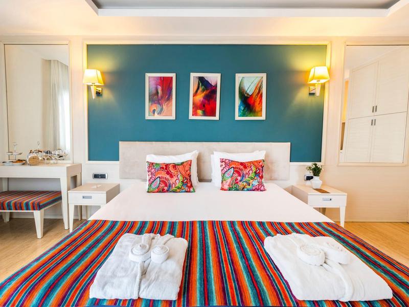 Chambre d'hôtel lumineuse avec lit double, coussins colorés et jeté de lit rayé, tables de chevet et tableaux muraux.