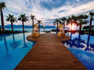 Wooden walkway leads to a pool with palm trees and a sunset view over the sea.